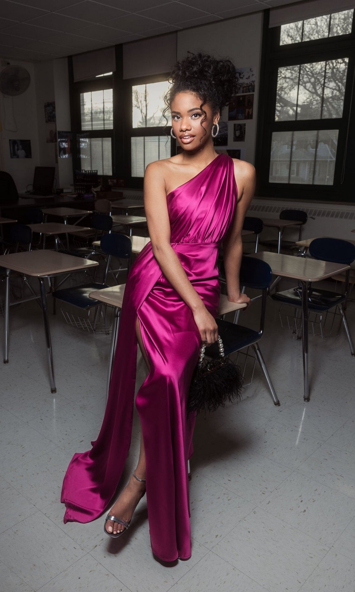 In a classroom, a person wears the Mac Duggal 26654 one-shoulder long formal dress in magenta, paired with silver heels and holding a small black handbag.